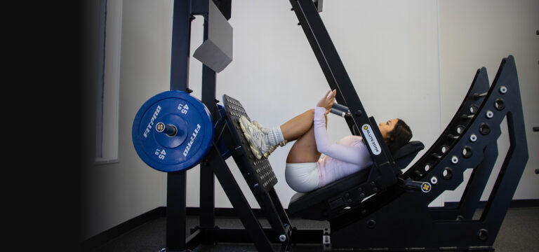 Woman training on a GluteZone Pendulum Squat machine in a bright studio, deep position with blue 45 lb plates.