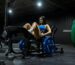 Mujer usando una maquina comercial de hip thrust con discos en un gimnasio oscuro.