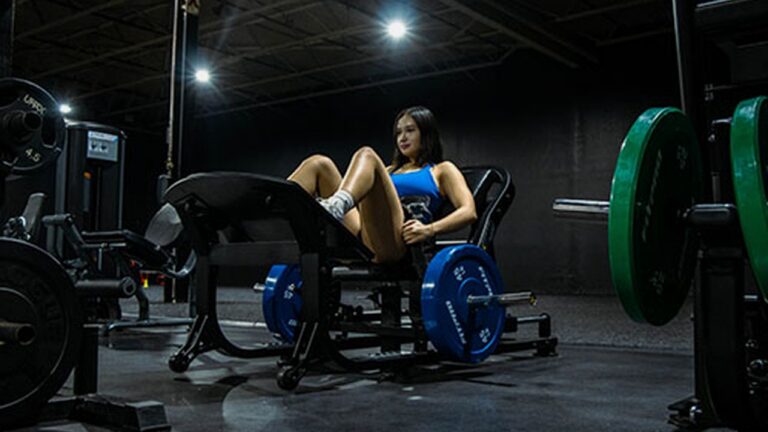 Mujer usando una maquina comercial de hip thrust con discos en un gimnasio oscuro.
