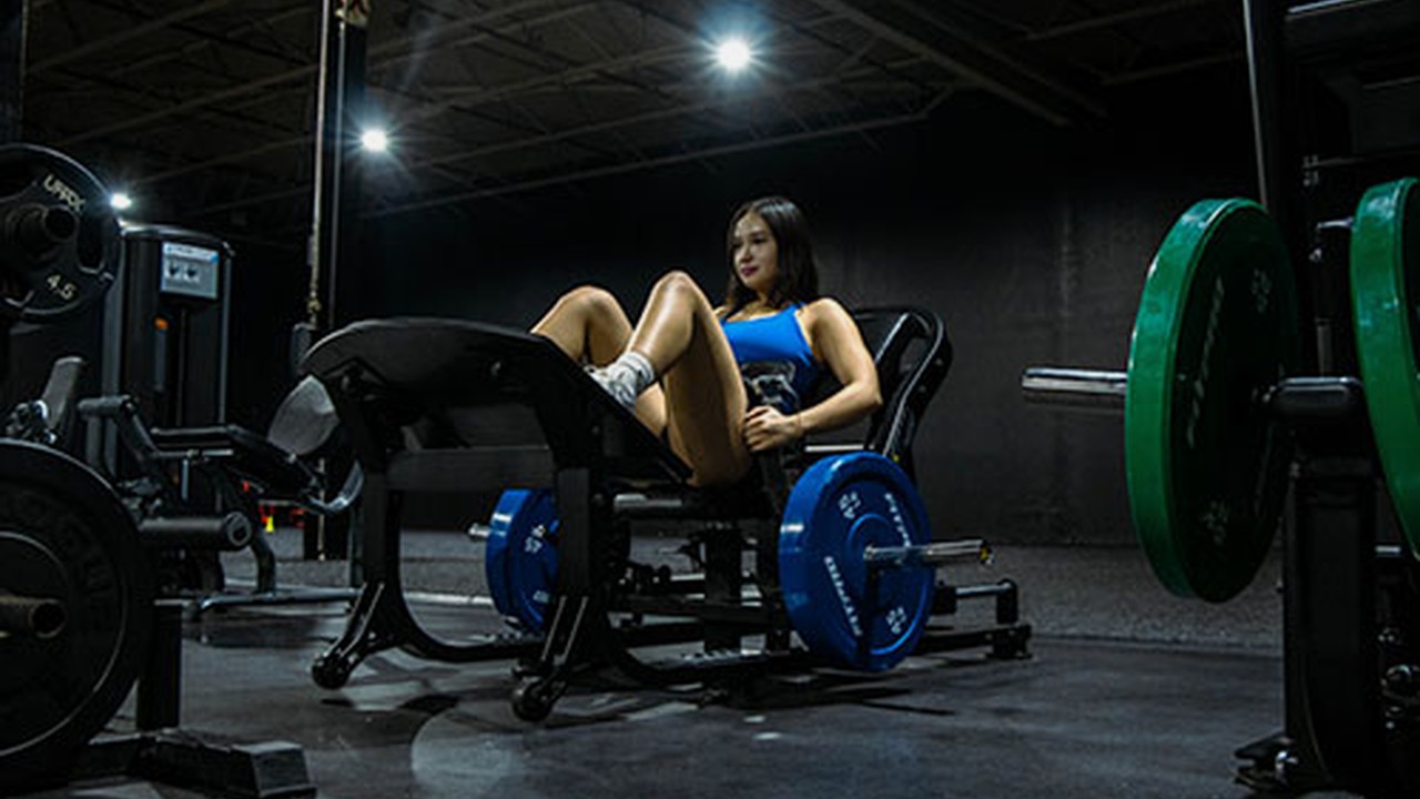 Mujer usando una maquina comercial de hip thrust con discos en un gimnasio oscuro.